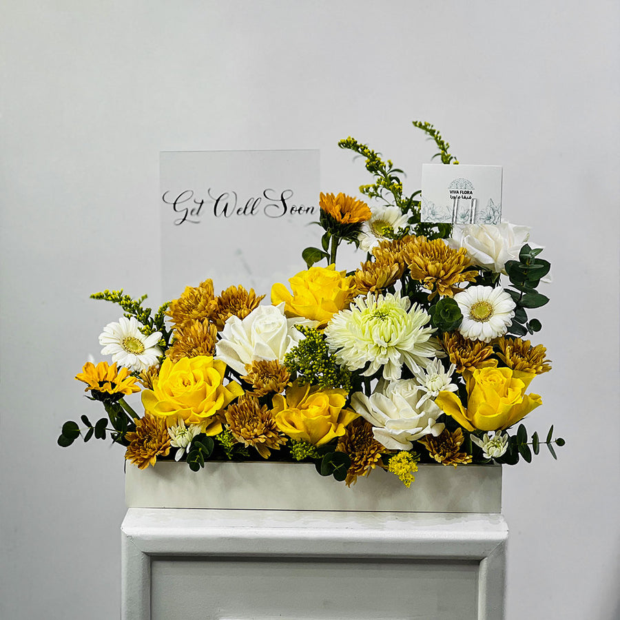 A rectangular white floral arrangement box filled with yellow and white flowers, including roses, chrysanthemums, and daisies, with green foliage. Two greeting cards are propped behind the arrangement against a light gray wall.