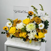 A floral arrangement in a white rectangular box on a white pedestal, featuring yellow and white roses, chrysanthemums, daisies, sunflowers, and green foliage, with a sign reading 'Get Well Soon' behind it.