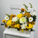 A floral arrangement in a white rectangular box on a white pedestal, featuring yellow and white roses, chrysanthemums, daisies, sunflowers, and green foliage, with a sign reading 'Get Well Soon' behind it.