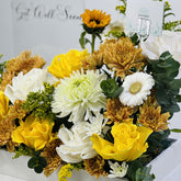 A floral arrangement in a white box featuring yellow and white roses, white and tan chrysanthemums, white daisies, green eucalyptus leaves, and small green filler flowers, with a small Viva Flora Greeting card.