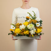 A rectangular white floral arrangement box filled with yellow and white flowers, including roses, chrysanthemums, and daisies, with green foliage. Two greeting cards are propped behind the arrangement against a light gray wall.