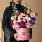 A pink floral arrangement in a pink bucket-style box with a handle, placed on a white pedestal against a pale gray background. The bouquet includes pink and purple flowers, green foliage, a small diploma prop, and a black-and-white graduation cap illustration insert.