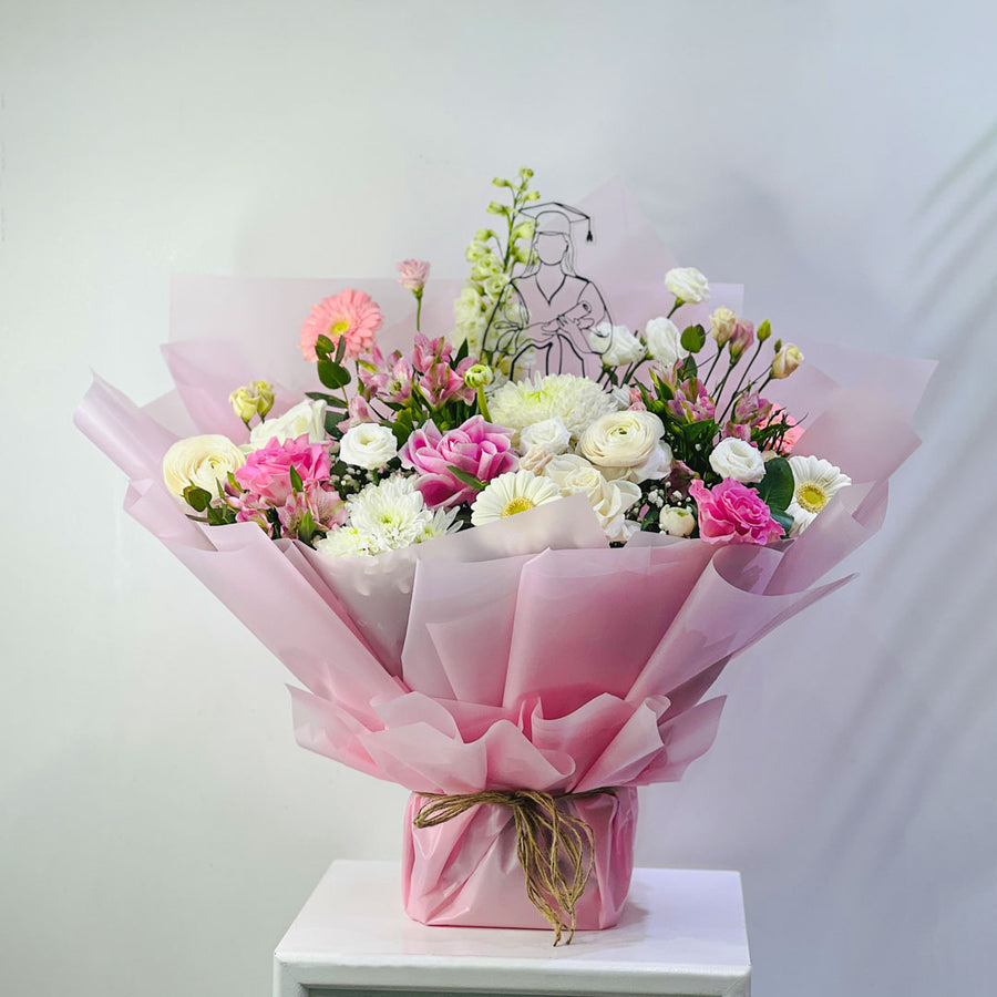 A vibrant floral bouquet wrapped in pink tissue paper and tied with a brown twine bow, displayed on a white pedestal against a light gray background.