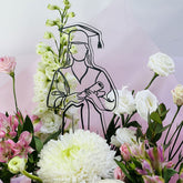 A graduation-themed floral arrangement with white and pink flowers surrounding a black line-art silhouette of a graduate holding a diploma, set against a soft pink and white background.