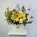 A graduation-themed floral arrangement in a white box with the brand name 'VIVA FLORA'. The bouquet features yellow roses, sunflowers, white carnations, green foliage, and a black wire graduation cap silhouette decoration, displayed on a white side table against a light gray wall.