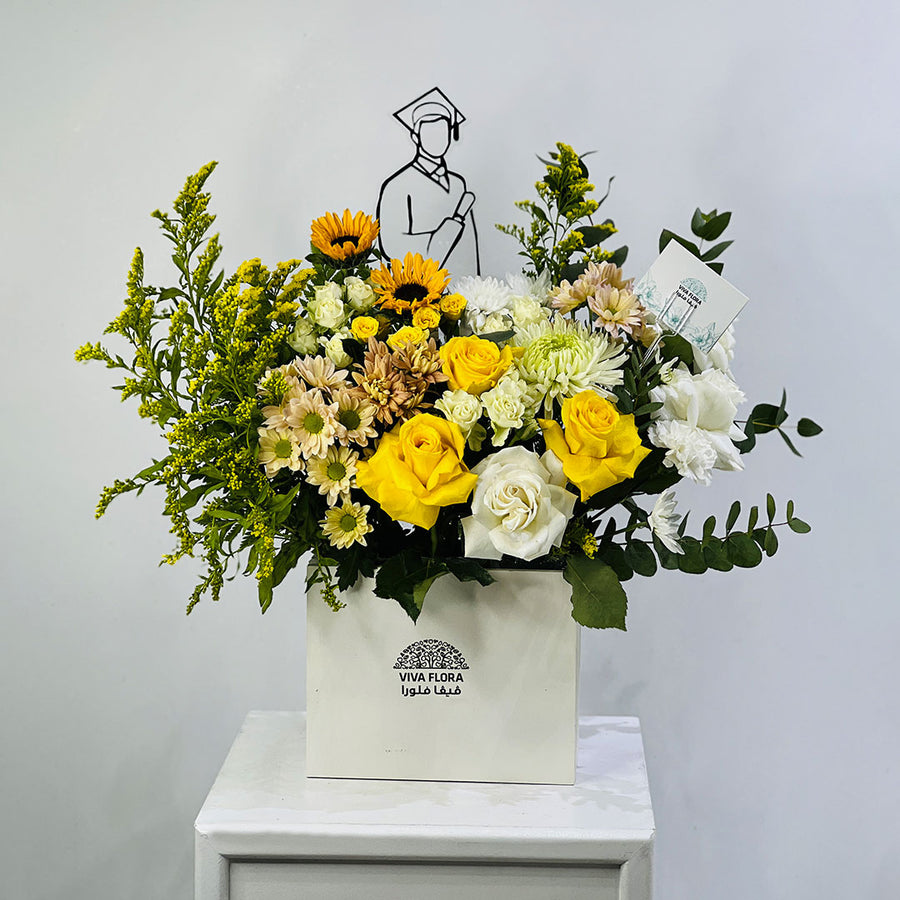 A graduation-themed floral arrangement in a white box with the brand name 'VIVA FLORA'. The bouquet features yellow roses, sunflowers, white carnations, green foliage, and a black wire graduation cap silhouette decoration, displayed on a white side table against a light gray wall.