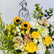 A vibrant graduation flower bouquet featuring yellow sunflowers, yellow and white roses, white and pale yellow daisies, green eucalyptus, and yellow bird's eye flowers, arranged in front of a white background with a simple black line drawing of a graduate.