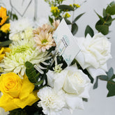 A close-up of a floral arrangement featuring white roses, white carnations, yellow roses, pale peach daisies, and green foliage, with a small printed card held in a clear stand.