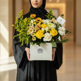 A graduation-themed floral arrangement in a white box with the brand name 'VIVA FLORA'. The bouquet features yellow roses, sunflowers, white carnations, green foliage, and a black wire graduation cap silhouette decoration, displayed on a white side table against a light gray wall.