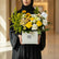 A graduation-themed floral arrangement in a white box with the brand name 'VIVA FLORA'. The bouquet features yellow roses, sunflowers, white carnations, green foliage, and a black wire graduation cap silhouette decoration, displayed on a white side table against a light gray wall.