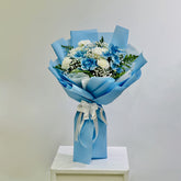 A bouquet of blue and white flowers wrapped in light blue paper and tied with a white ribbon, displayed on a white pedestal against a light gray background.