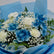 A floral bouquet featuring blue and white flowers, including carnations, roses, and baby's breath, wrapped in light blue tissue paper.