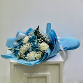 A bouquet of blue and white flowers wrapped in light blue paper, placed on top of a white pedestal against a plain light gray background.