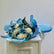 A bouquet of blue and white flowers wrapped in light blue paper, placed on top of a white pedestal against a plain light gray background.