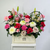 A vibrant floral arrangement in a cream-colored rectangular box on a white pedestal, featuring pink, red, white, and green flowers with a gold 'Happy Birthday' sign, from the brand VIVA FLORA.