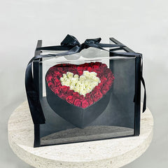Heart-shaped arrangement of red and white roses in a clear box with a black ribbon on a light gray background.
