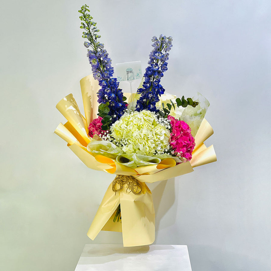 A wrapped flower bouquet featuring tall blue delphiniums, a central cluster of pale yellow hydrangeas, bright pink carnations, and green foliage, wrapped in shiny gold paper and tied with a gold ribbon, displayed on a white pedestal against a light gray background.