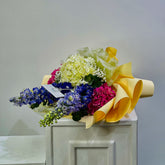 A multicolor floral arrangement featuring blue delphiniums, pink carnations, and a pale yellow hydrangea, wrapped in yellow paper and displayed on a white pedestal against a plain gray wall.