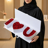 A white gift box filled with red roses arranged to spell out 'I <heart> U' on a light stone surface against a warm beige background.