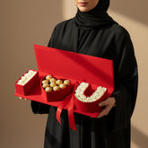 A red gift box shaped like the word 'I LOVE U' filled with chocolates, displayed on a white marble pedestal against a warm beige background. The letters 'I' and 'U' are filled with small white flowers, the 'O' is filled with Ferrero Rocher-style chocolates wrapped in gold foil, and a red ribbon sits in front of the box.