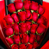A bouquet of red roses wrapped in bright red and black paper, held by a hand against a light marble background.