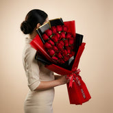 A large bouquet of red roses wrapped in bright red and black paper, held by a person's arm on the left. The bouquet is tied with a red ribbon printed with 'Viva Flora'.
