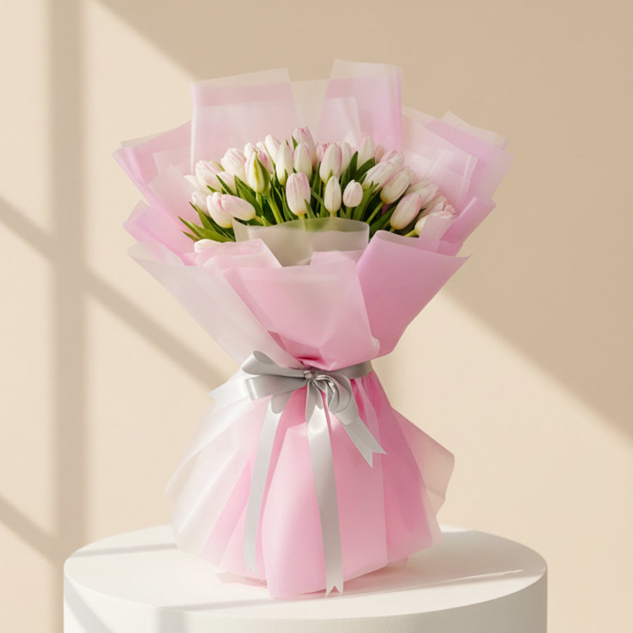 A bouquet of white tulips wrapped in pink tissue paper and tied with a silver ribbon, held by a person whose arm is visible on the left side, set against a marble-patterned wall background.