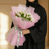 A bouquet of white tulips wrapped in pink tissue paper and tied with a silver ribbon, held by a person whose arm is visible on the left side, set against a marble-patterned wall background.