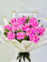 A bouquet of pink roses wrapped in white paper with gold trim and tied with a black ribbon, photographed against a plain light background.