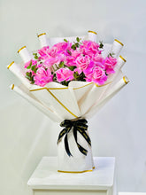 A bouquet of pink roses wrapped in white paper with gold trim and tied with a black ribbon, displayed on a white pedestal against a light gray background.