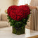 A heart-shaped bouquet of red roses arranged in a green basket with a black ribbon, displayed on a white pedestal against a plain light background.