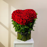 A heart-shaped bouquet of red roses arranged in a cylindrical vase with green foliage and a black ribbon, displayed on a white pedestal against a plain light background.