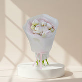 A bouquet of pale pink and white peony flowers wrapped in translucent white paper with a pink tissue lining and pink ribbon, photographed against a soft light gray background.