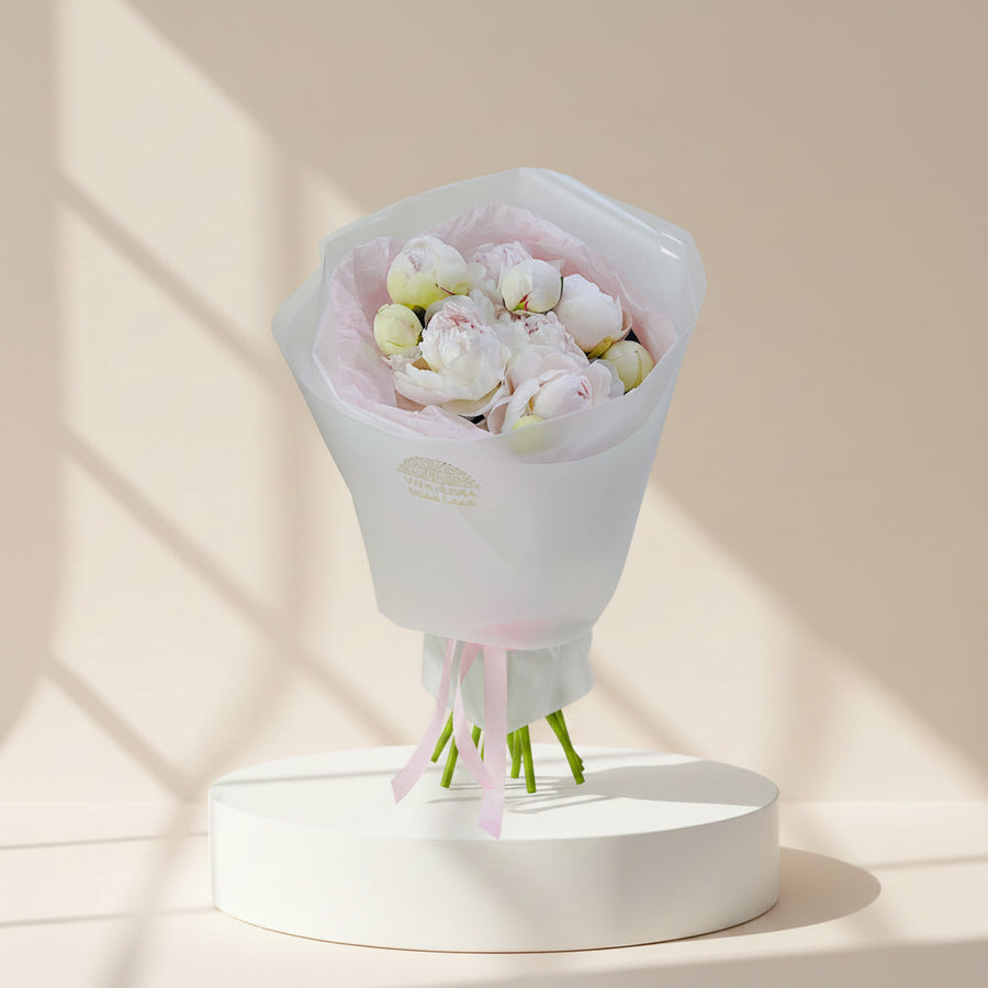 A bouquet of pale pink and white peony flowers wrapped in translucent white paper with a pink tissue lining and pink ribbon, photographed against a soft light gray background.