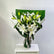 A bouquet of white lilies with green leaves and buds, wrapped in green paper and a white paper sleeve with a silver ribbon, displayed on a white pedestal against a light gray background.