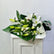 A bouquet of white lilies with green leaves and buds, wrapped in dark green paper with a white ribbon, resting on a white cabinet against a light gray wall.