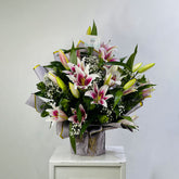 A large floral arrangement of pink and white lilies with green leaves and small white baby's breath, wrapped in gray paper and displayed on a white pedestal against a light gray wall.