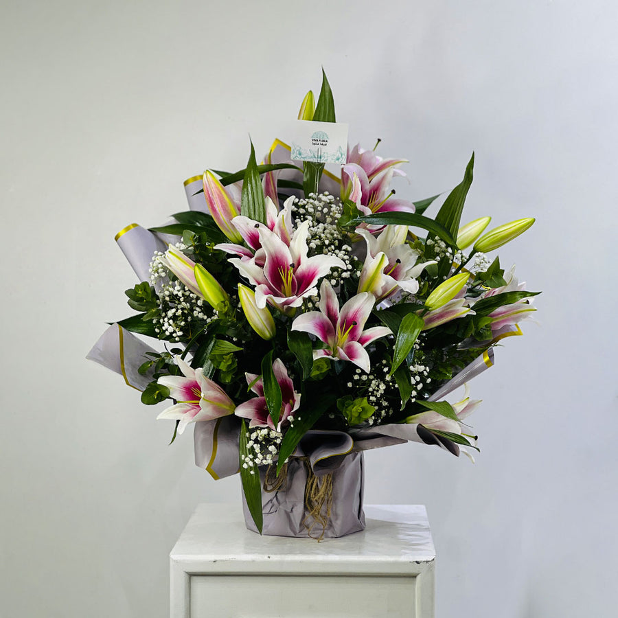 A large floral arrangement of pink and white lilies with green leaves and small white baby's breath, wrapped in gray paper and displayed on a white pedestal against a light gray wall.