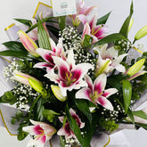 A vibrant bouquet of lilies and baby's breath flowers wrapped in white and gold paper, photographed from a top-down angle against a clean white background.