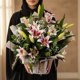 A large floral arrangement of pink and white lilies with green leaves and small white baby's breath, wrapped in gray paper and displayed on a white pedestal against a light gray wall.