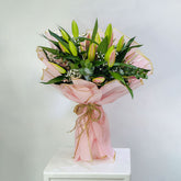 A fresh flower bouquet wrapped in light pink tissue paper and tied with a rustic jute twine bow, displayed on a white pedestal against a neutral light gray background.