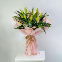 A fresh flower bouquet wrapped in light pink tissue paper and tied with a rustic jute twine bow, displayed on a white pedestal against a neutral light gray background.