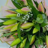 A close-up of a flower bouquet featuring pink and green lily buds, lush green leaves, and small white baby's breath flowers wrapped in pale pink paper.