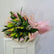 A fresh flower bouquet with green lily buds, pink lilies, green foliage, and small white baby's breath, wrapped in pink tissue paper with gold foil edges and tied with twine, displayed on a white cabinet against a light gray wall.