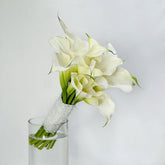 A bouquet of white calla lilies with green stems wrapped in white lace, displayed in a clear glass vase against a light gray background.