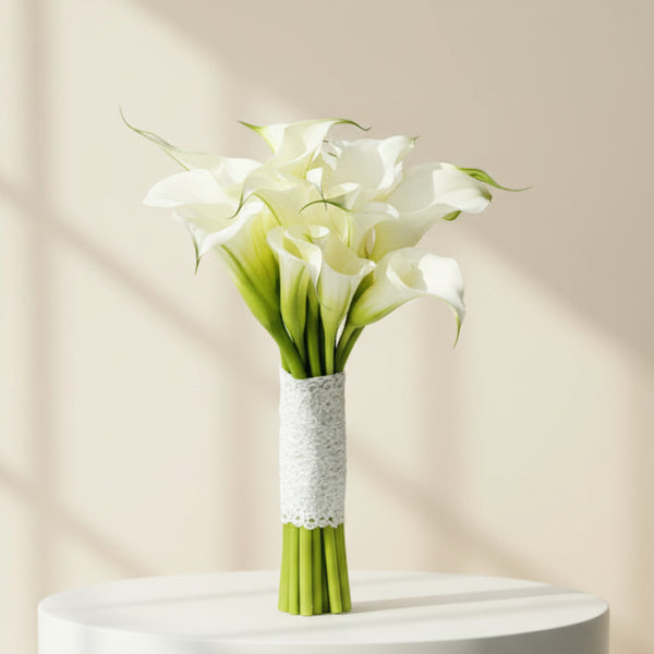 A bouquet of white calla lilies with green stems wrapped in white lace, displayed in a clear glass vase against a light gray background.