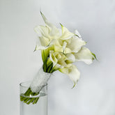 A bouquet of white calla lilies with green stems, wrapped in a white lace ribbon, arranged in a clear glass vase against a light gray background.