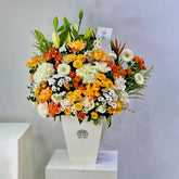 A vibrant floral arrangement featuring orange, yellow, and white flowers with green foliage, presented in a white geometric vase on a white pedestal against a light gray background.