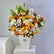 A vibrant floral arrangement featuring orange, yellow, and white flowers with green foliage, presented in a white geometric vase on a white pedestal against a light gray background.