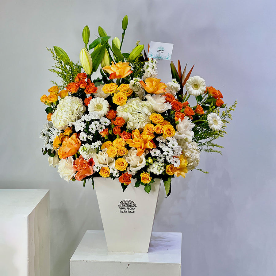 A vibrant floral arrangement featuring orange, yellow, and white flowers with green foliage, presented in a white geometric vase on a white pedestal against a light gray background.